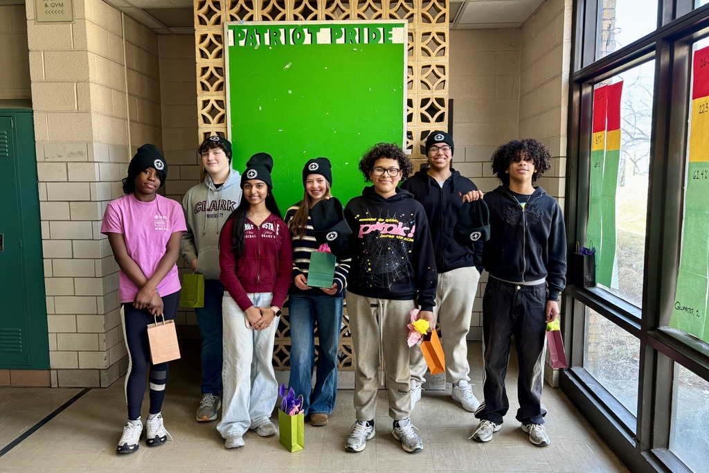 Seven students stand together in a hallway, holding gift bags and wearing beanies.