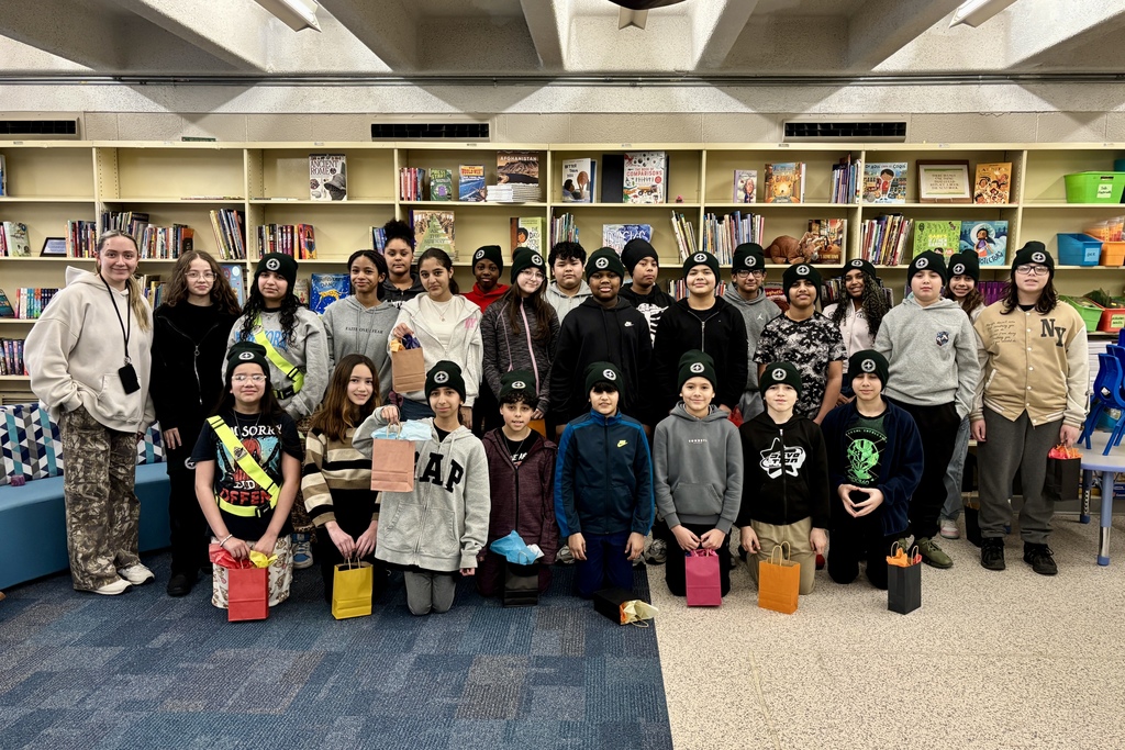 A group of students and an adult pose for a photo in front of bookshelves, many wearing matching beanies and holding gift bags.