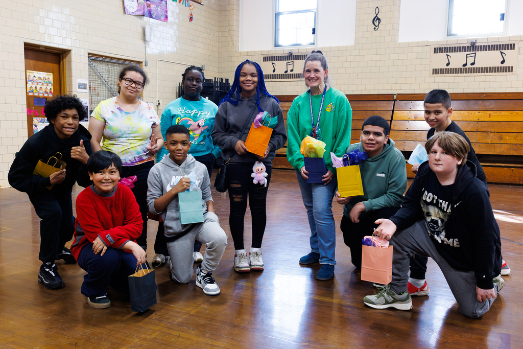 A group of students and their teracher stand in a gymnasium, holding colorful gift bags.