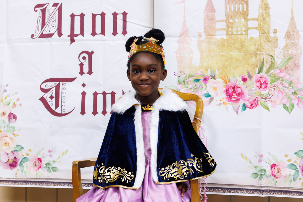 A young girl in a princess costume sits on a chair in front of a banner that reads 'Upon a Time' and features a castle and floral design.