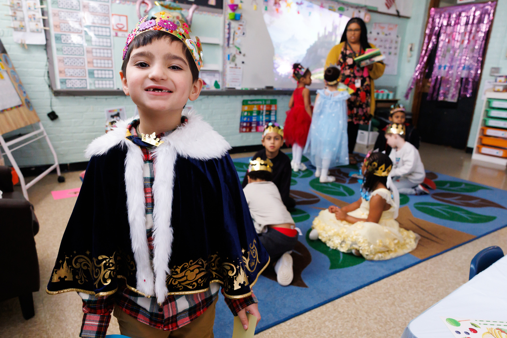 A young boy in a king's costume and crown smiles at the camera in a classroom.