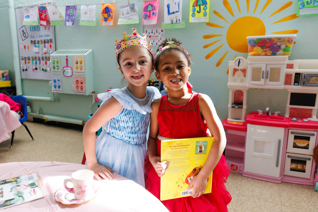 Two young girls, one in a blue princess dress and the other in a red dress, smile at the camera, holding a book titled 'The King of Kindergarten'.