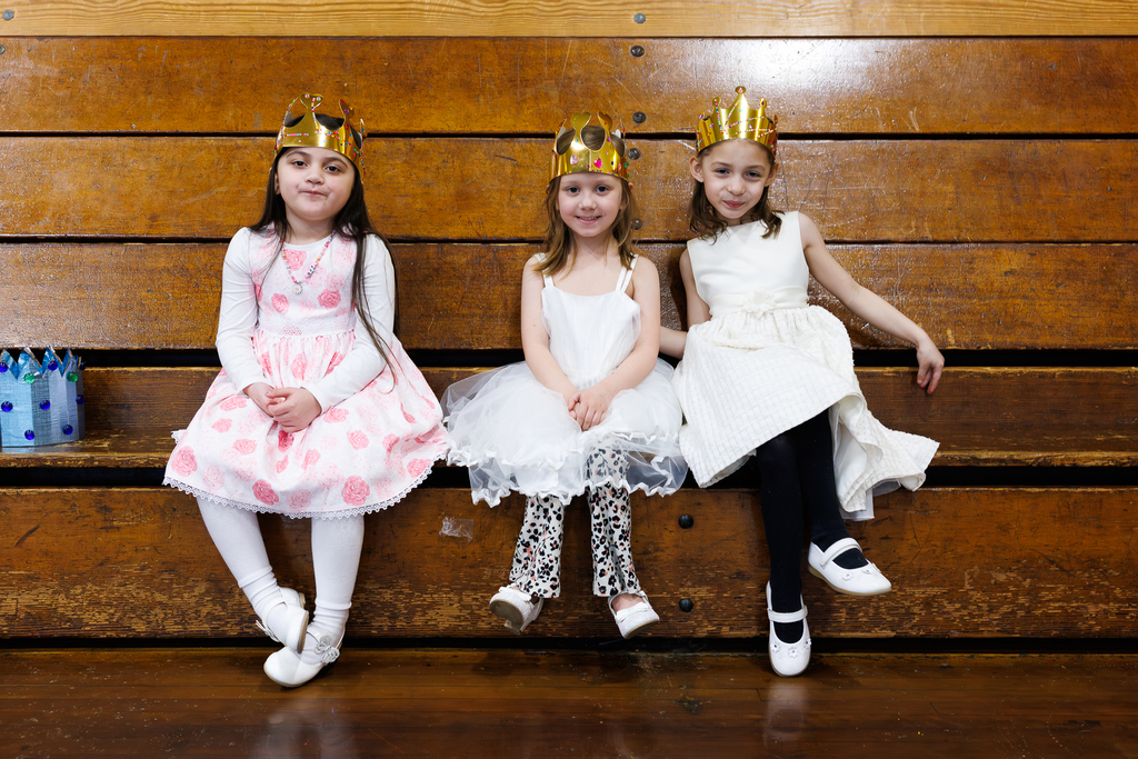Three young girls wearing golden crowns sit on a wooden bleacher, dressed in festive attire.