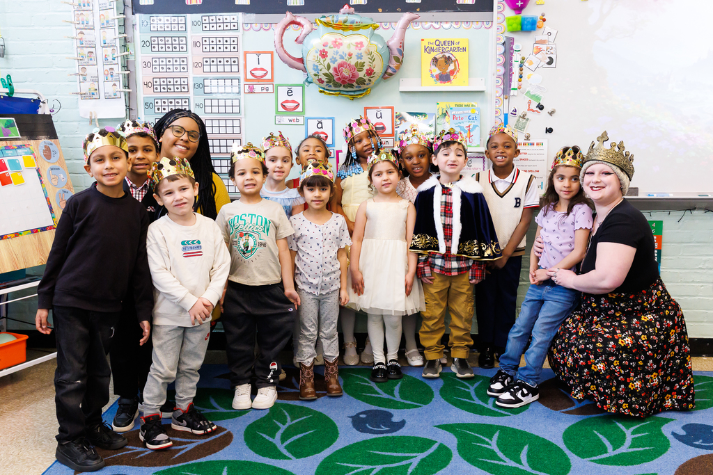A diverse group of kindergarten students and their teacher, all wearing crowns, pose for a photo in a classroom decorated with educational charts and a teapot balloon.