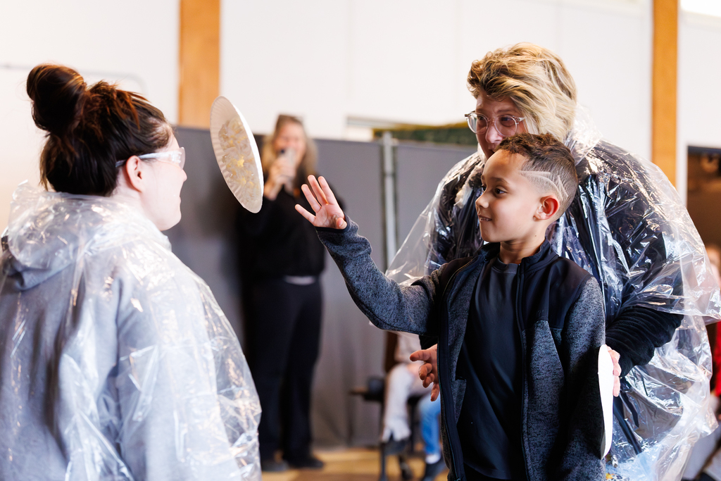 With the help of a teacher, a student throws a pie into the face of another teacher during a school assembly.