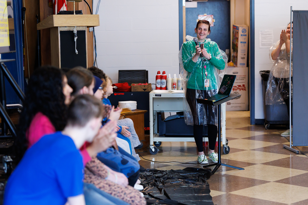 A teacher wearing a plastic poncho talks about a student during a school assembly.
