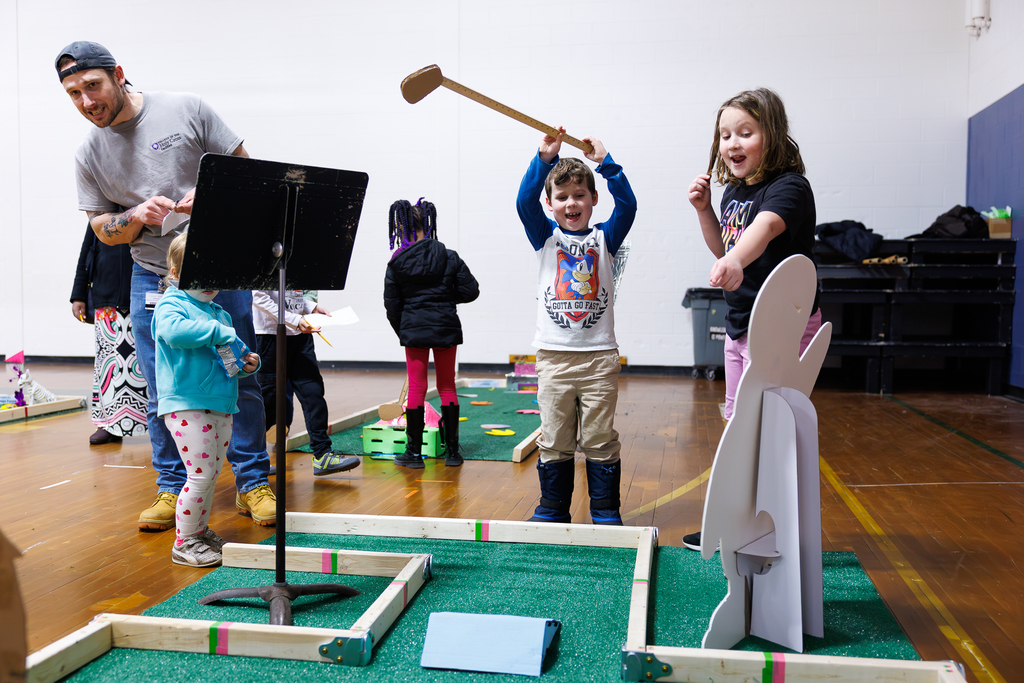 A mini-golfer throws their arms in the air to celebrate a putt while their parent and sibling look on and cheer.