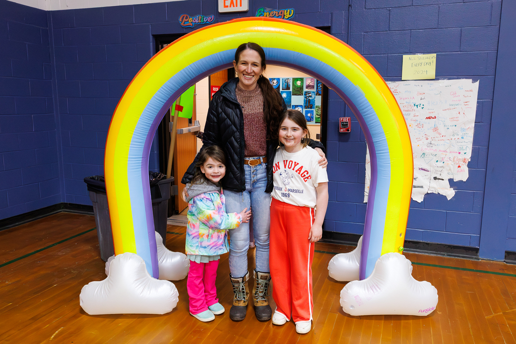 A parent and their two young children stand together for a photo underneath a rainbow archway.