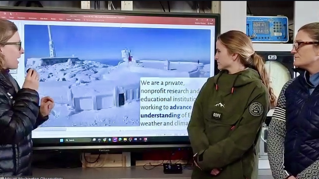 Three people stand in front of a large screen displaying a snowy mountain landscape and text about research.
