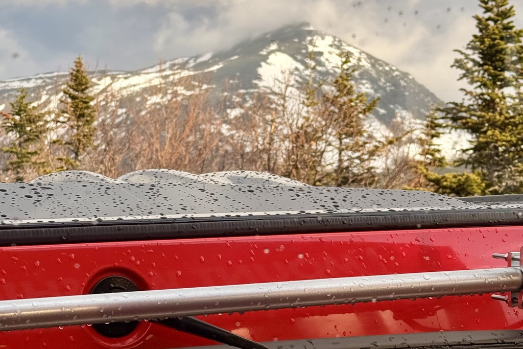 A red vehicle's surface is wet with raindrops, with a snow-capped mountain and pine trees in the background.
