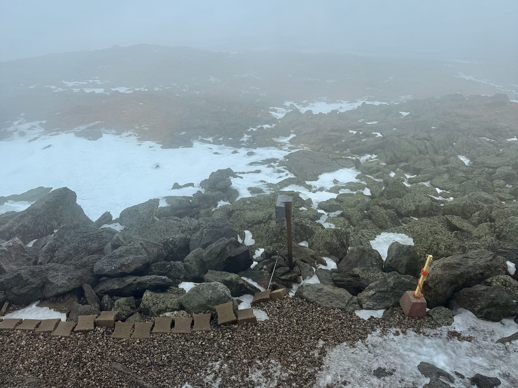 A rocky, misty mountain summit with patches of snow and a small marker.