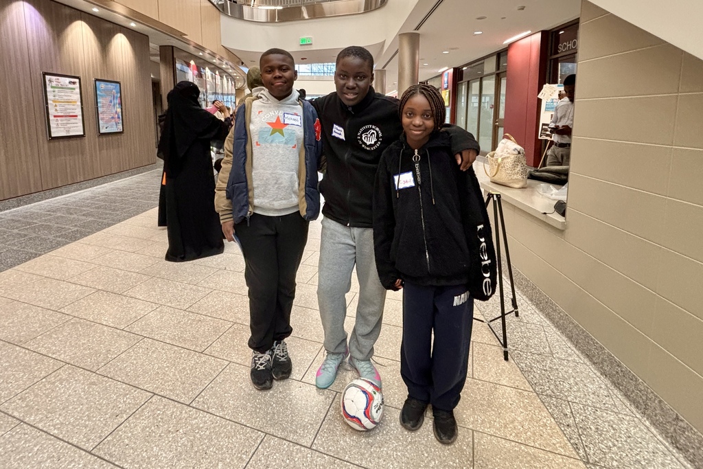 Three young people stand together in a hallway, with a soccer ball at their feet.