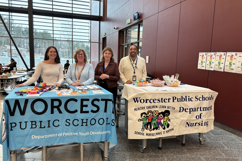 Four smiling individuals stand behind tables displaying information and promotional items for Worcester Public Schools.