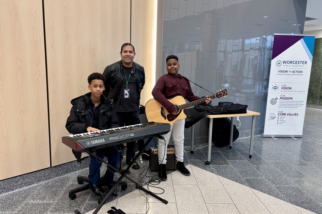 A young person plays a Yamaha MOXF6 keyboard while another strums an acoustic guitar, with an adult standing behind them.