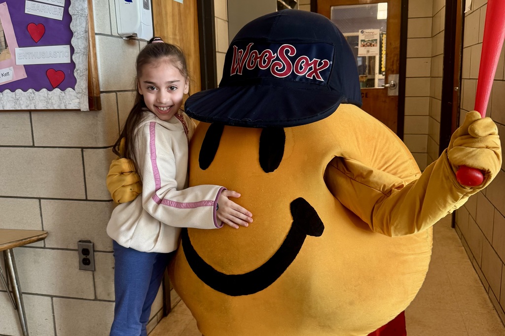 A young girl smiles as she hugs a mascot wearing a WooSox baseball cap.