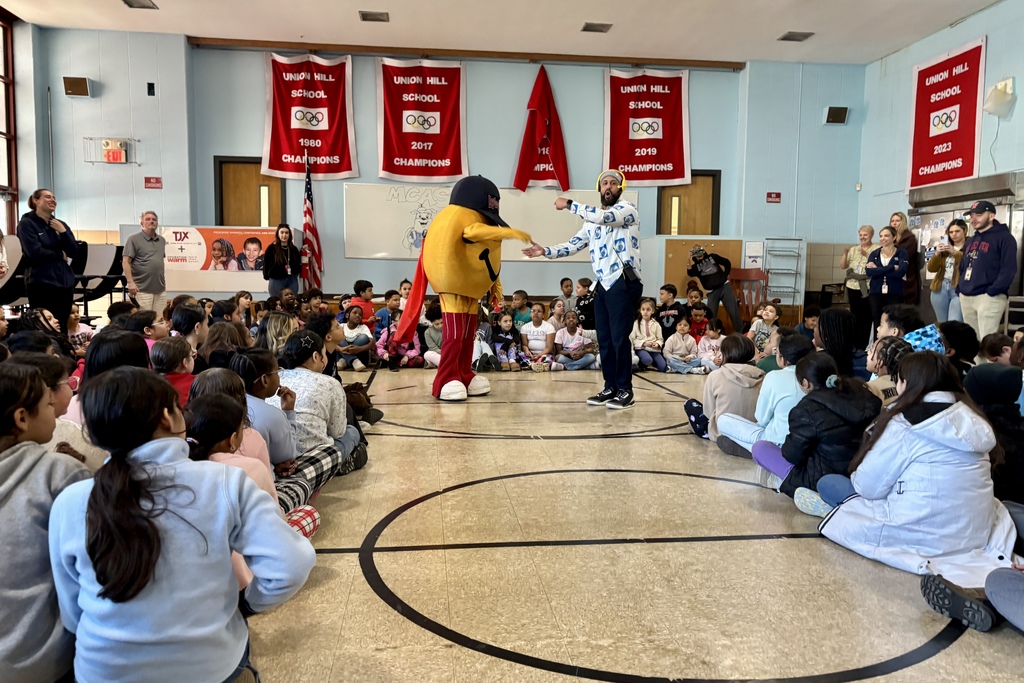 A school gymnasium filled with students sitting on the floor, watching a mascot and a presenter.