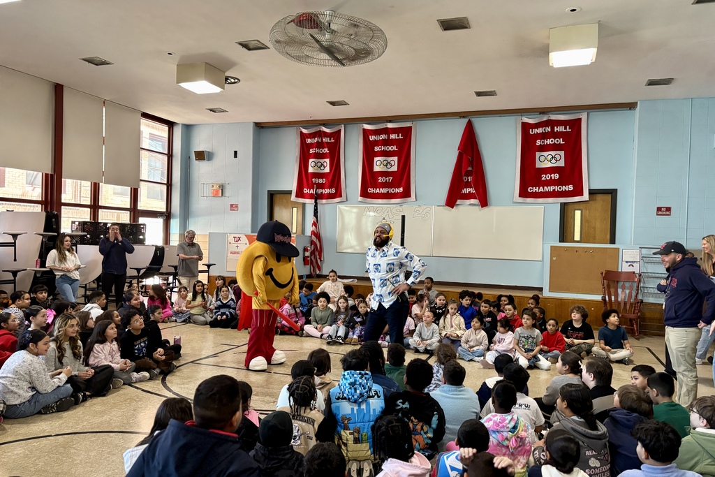 A school gymnasium filled with students sitting on the floor, watching a mascot and a presenter.