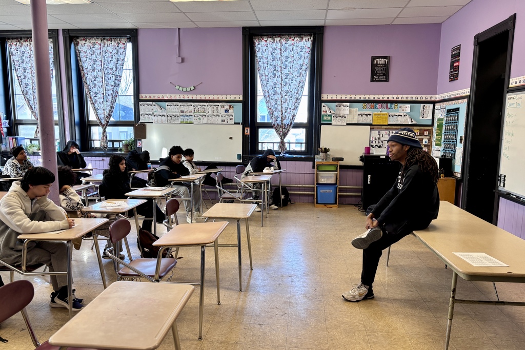 Students sit at desks in a classroom with purple walls and patterned curtains.