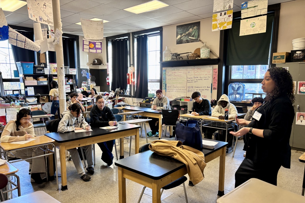 A teacher with a name tag that reads 'Coral Pacheco' gestures while speaking to students seated at desks in a classroom.