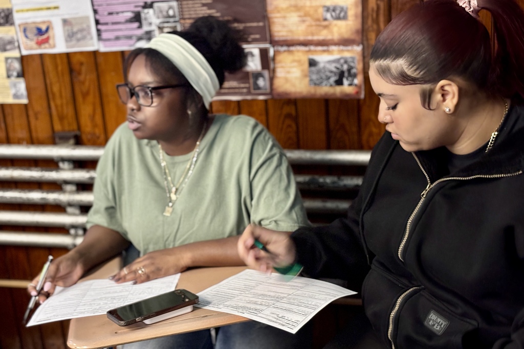 Two young women are seated at desks, focused on papers and writing.
