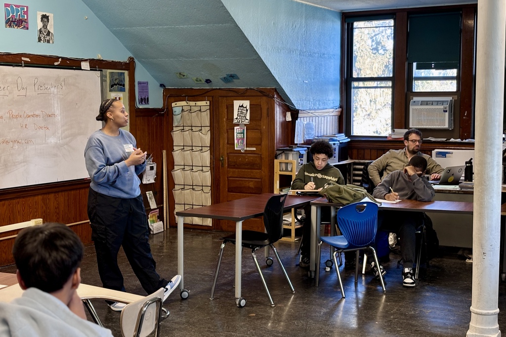 A presenter stands near a whiteboard in a classroom, addressing students seated at desks.