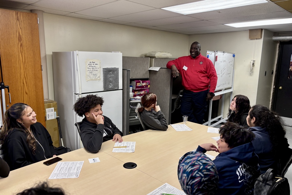 A diverse group of students and an adult in a red jacket are gathered around a table in a classroom.