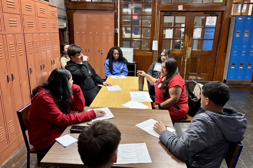 A group of students and an instructor sit around tables in a room with lockers, discussing papers.
