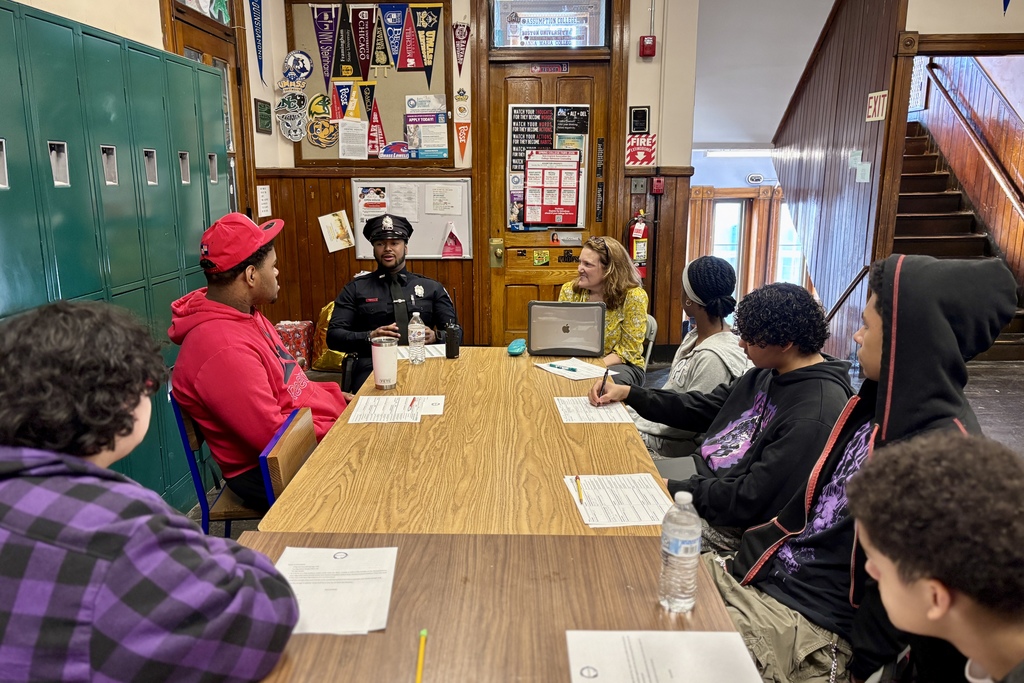 A police officer leads a discussion with students and a teacher around a table in a classroom with lockers.