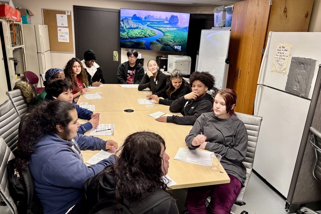 A diverse group of young people sit around a large table, engaged in a discussion or activity.