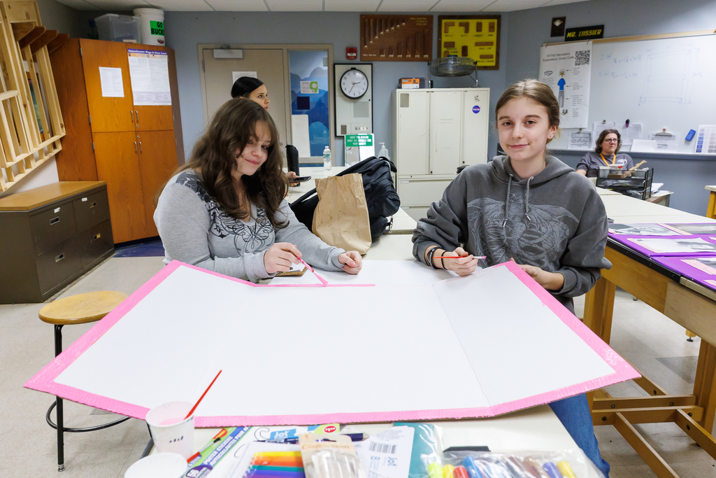 Two students work on a large white project with pink trim, using brushes and markers.