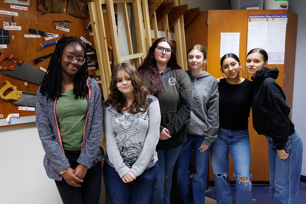 Six students stand together in a workshop, with tools and educational posters visible in the background.