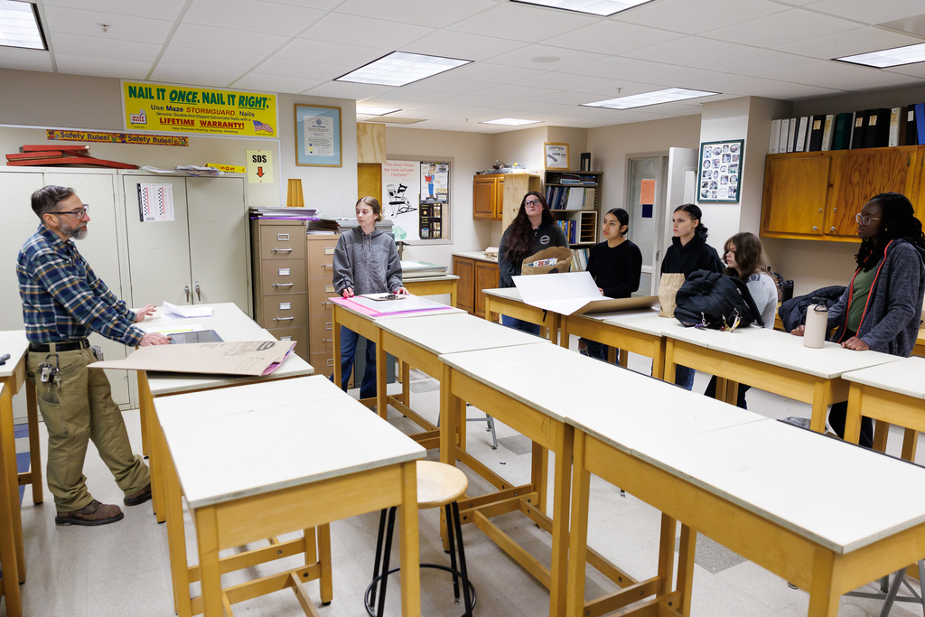 A teacher instructs a group of students in a classroom with drafting tables and supplies.
