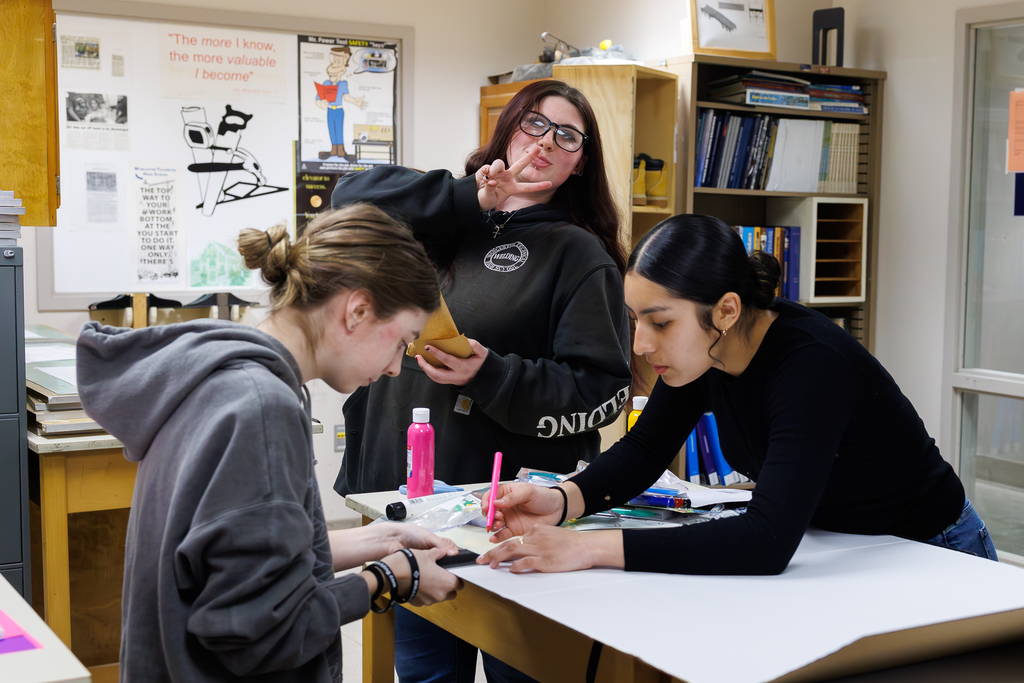 Three students collaborate on a project at a table, with one making a peace sign.