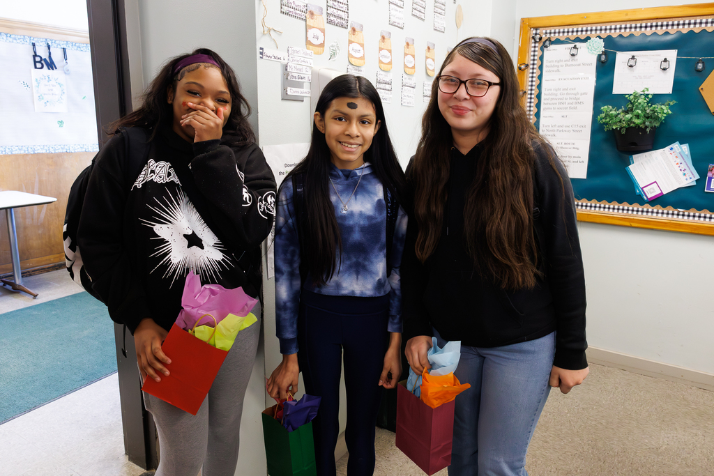 Three students stand together, each holding a gift bag filled with colorful tissue paper.