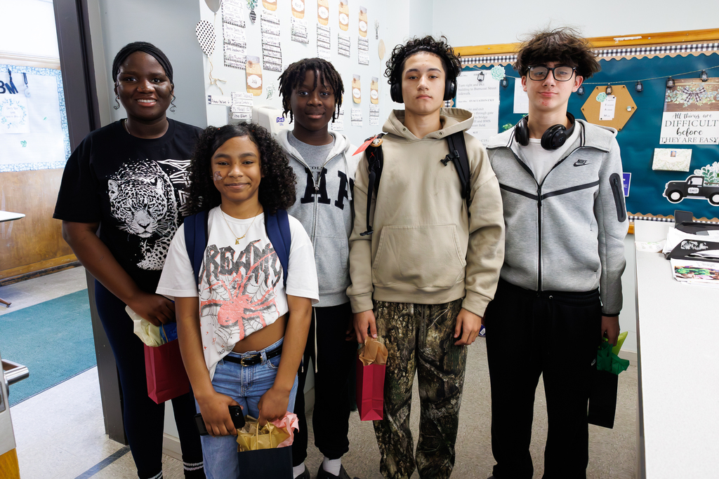 Five students stand together, each holding a gift bag filled with colorful tissue paper.