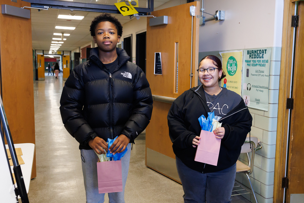 Two students stand together, each holding a gift bag filled with colorful tissue paper.
