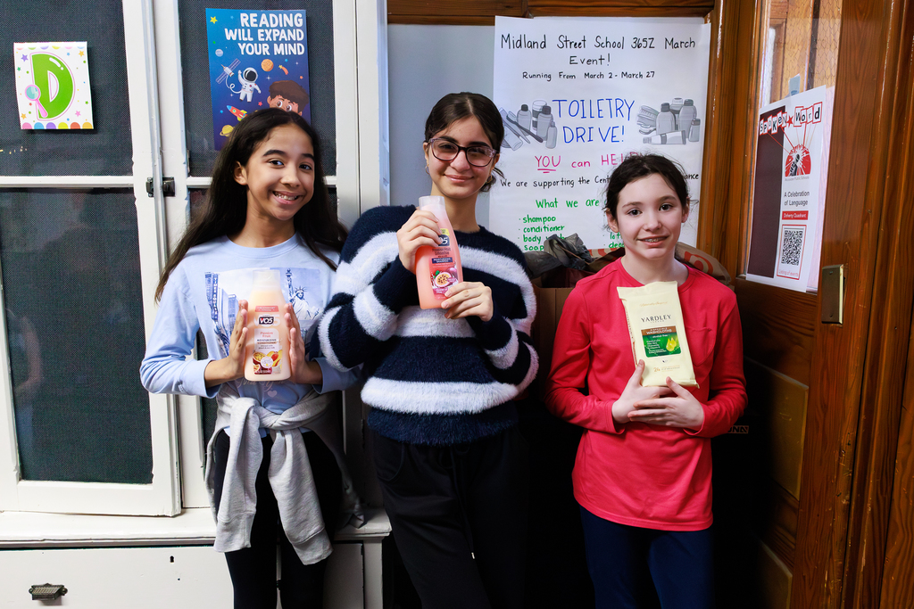 Three smiling students hold toiletries for a school drive, with posters about reading and the event in the background.