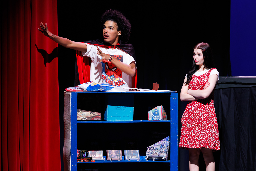 An actor in a Spider-Man shirt and cape gestures dramatically on stage, with a young woman in a red floral dress standing beside him.