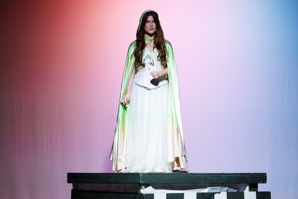 An actor in a white gown and a flowing green cape stands on a stage.