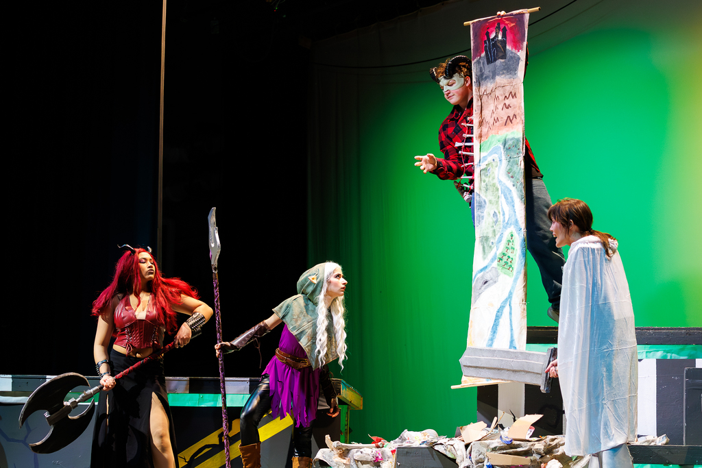 Actors in fantasy costumes perform on a stage with a green backdrop.