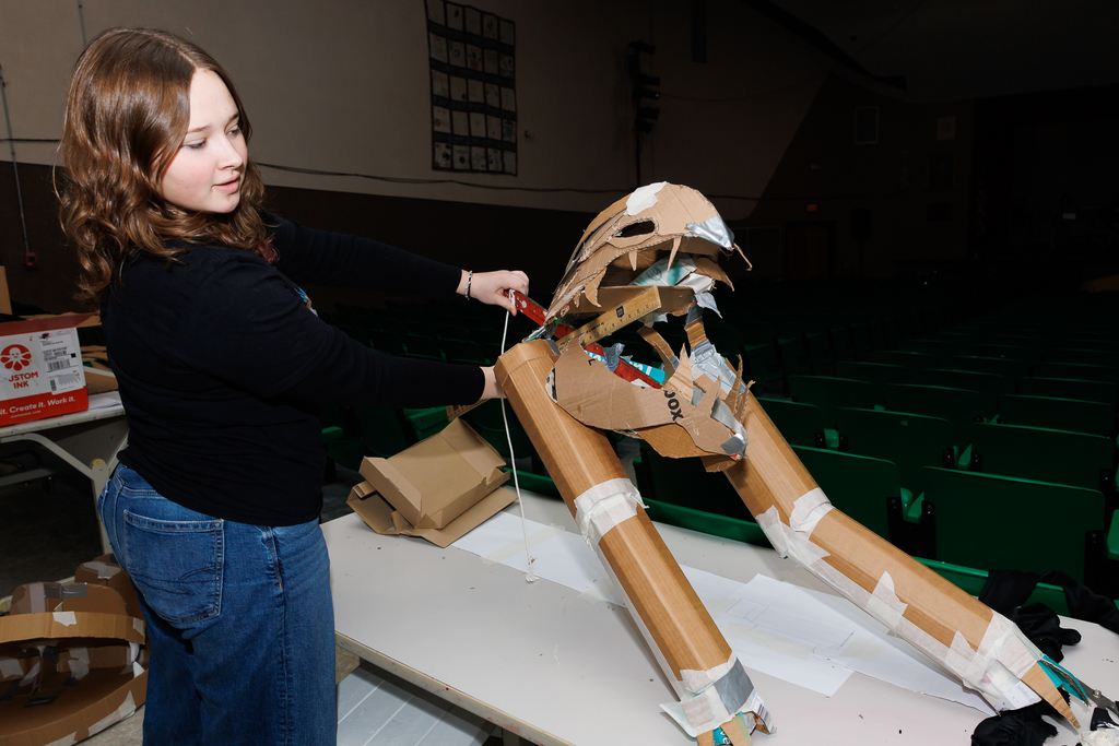 A student works on a large cardboard sculpture, using a ruler and scissors.