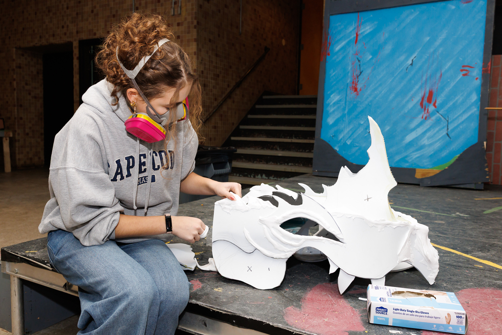 An actor wearing a respirator mask and a grey hoodie works on a large, white, dragon-like prop.