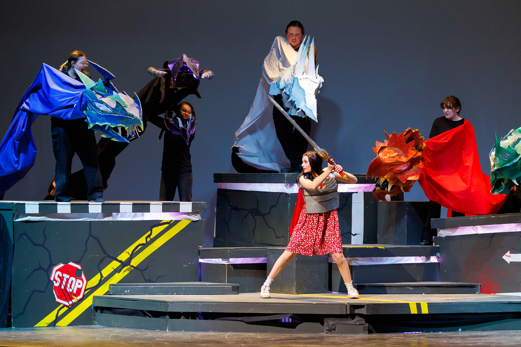 An actor in a red dress and armor wields a sword on a stage with dragon puppets.