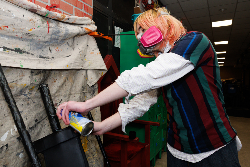 A student wearing a pink respirator mask sprays a can of black paint onto a prop.