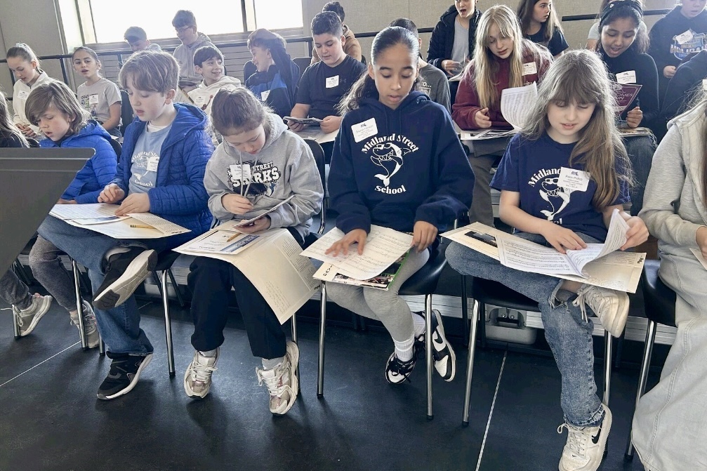 A group of children sit in chairs, looking at papers and engaging in an activity.