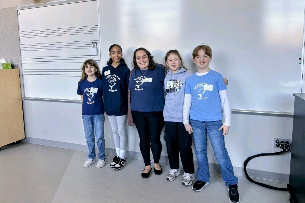 A group of five students and one adult stand in front of a whiteboard with musical staff lines.