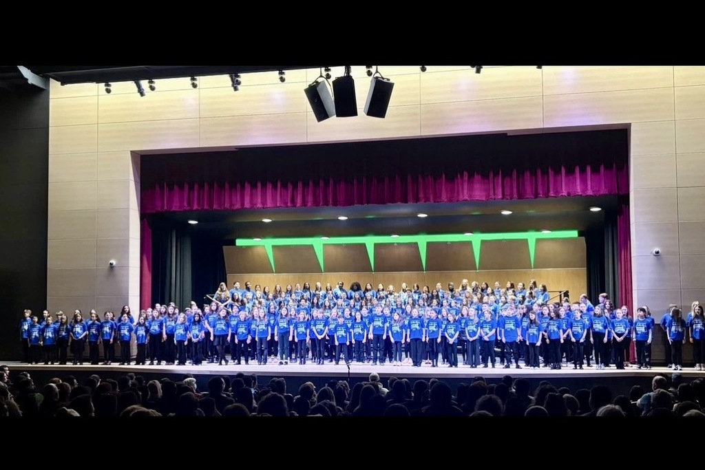 A large choir of students in blue t-shirts stands on a stage in front of a purple curtain.