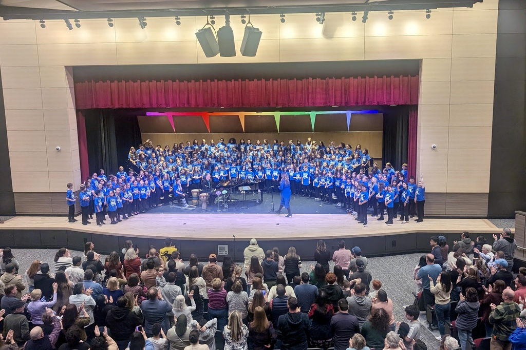 A large choir of students in blue shirts stands on a stage, facing an audience.