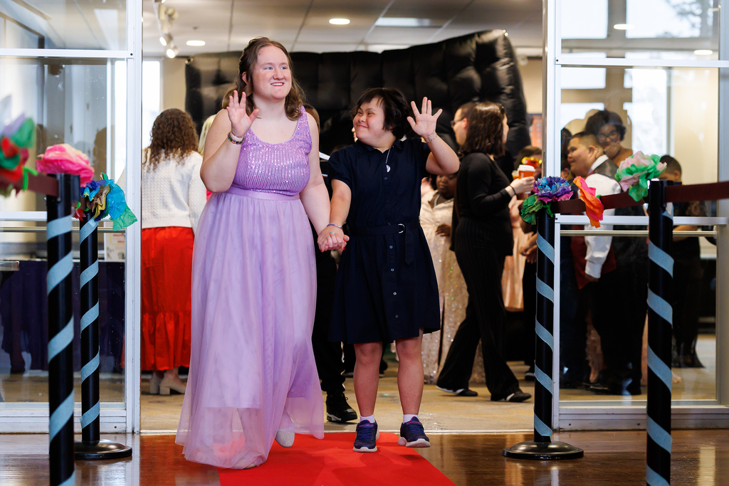 Two students walk down the red carpet while holding hands, waving at the crowd of spectators.