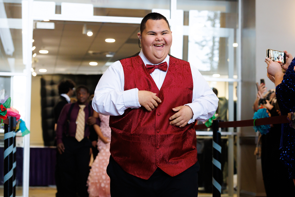 A student in a white shirt and red vest smiles broadly while walking.